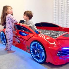 two children are playing in a red car bed with lights on the sides and blue wheels