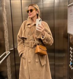 a woman taking a selfie in front of an elevator door wearing a tan coat