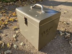 an old metal box with writing on it sitting in the middle of a sidewalk next to leaves