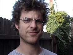 a man with curly hair wearing glasses in front of a wooden fence and green bushes