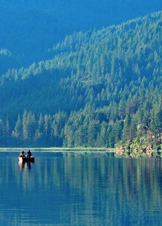 two people on a boat in the middle of a lake surrounded by mountains and trees