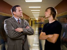 two men standing in an office hallway with their arms crossed and looking at each other