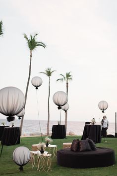 an outdoor setting with round tables and lanterns on the grass by the water in front of palm trees