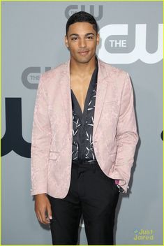 a man in a pink jacket and black shirt standing on a red carpet with his hands in his pockets