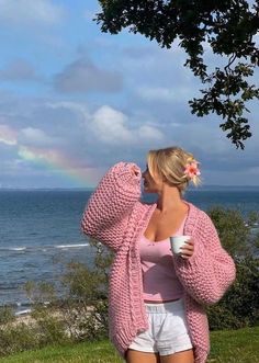 a woman standing on top of a lush green hillside next to the ocean holding a cup