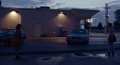 two people standing in front of a building at night with their cars parked on the street