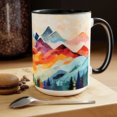 a coffee mug with mountains painted on it and a spoon sitting next to the cup