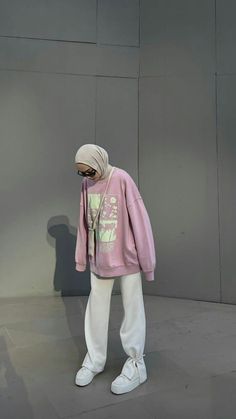 a woman in a pink sweatshirt and white pants stands against a gray wall with her hands on her hips