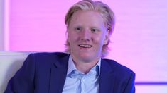 a man with blonde hair wearing a blue jacket and striped shirt sitting in front of a pink background