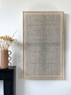 a wall hanging on the side of a white wall with a vase and plant in it