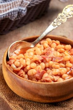 a wooden bowl filled with beans and bacon on top of a table next to a spoon