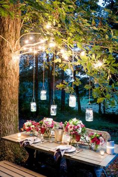 a table with flowers and candles on it under some lights hanging from the tree branches