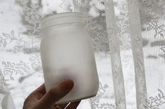 a hand holding a glass jar in front of a window with white lace on it