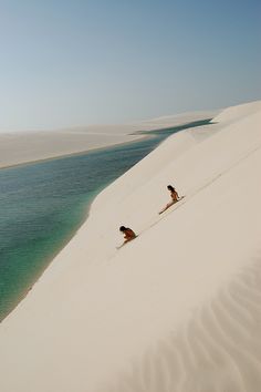 two people sitting on top of a white sand dune next to the ocean and water