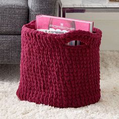 a red knitted basket with magazines in it on the floor next to a couch