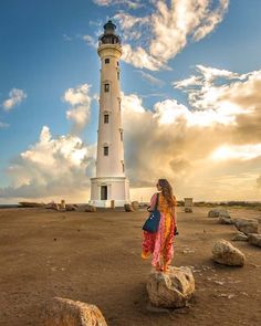 a woman standing in front of a light house on the beach with rocks around her