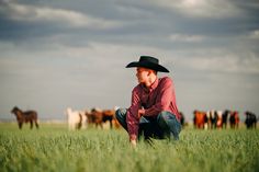 a man wearing a cowboy hat kneeling in the grass with cows behind him on a cloudy day