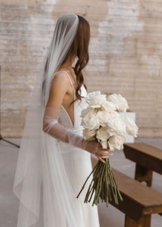 a woman in a white wedding dress holding a bouquet of flowers next to a bench