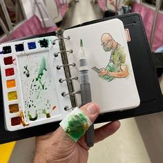 a hand holding an open notebook with watercolors on it and a drawing book in the background