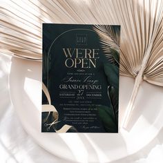 a black and gold wedding card on a plate next to some palm leaves, with the words we're open above it