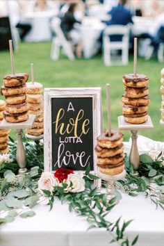 A wedding tables cape with greenery features a chalkboard sign with "A whole lotta love" written on it. Also on the table are several pedestals with multiple donuts on each pedestal, ready for guests to enjoy. Wedding Donut Dessert Table, Donut Dessert Table, Bridal Brunch Decorations, Donut Bar Wedding, Outdoor Wedding Backdrops, Donut Display, Wedding Donuts, Donut Dessert