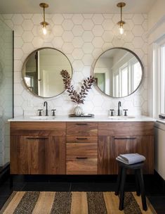 two round mirrors are above the double sinks in this bathroom with wood cabinetry and white hexagonal tiles