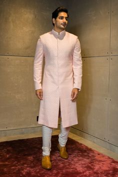 a man in a pink sherwar stands on a red carpet and poses for the camera