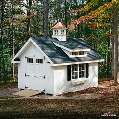 a small white shed with a steeple on the roof and windows is surrounded by trees