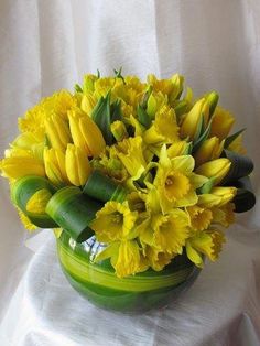 a green vase filled with yellow flowers on top of a white cloth covered tablecloth