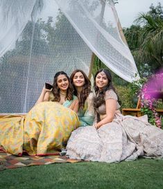 three women are sitting on the grass under a white net and posing for a photo