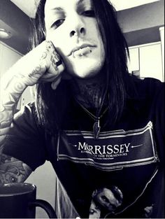 a man with long hair and piercings sitting in front of a coffee cup holding his hand to his face