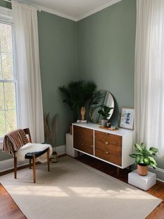 a living room with green walls and white drapes on the windowsill, a wooden sideboard, potted plants and a round mirror