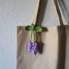 two crocheted purple flowers hang from a tan tote bag hanging on a wall