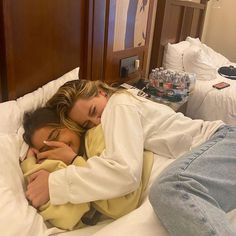 two women are cuddling on a bed with white sheets and blue jeans, one is wearing a yellow sweater