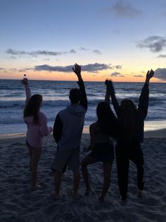 four people standing on the beach with their arms in the air, watching the sun set