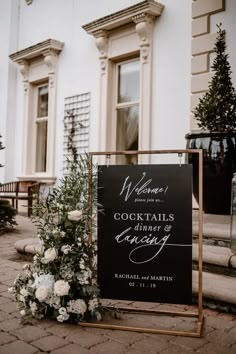 a sign that is sitting in front of a building with flowers and greenery on it