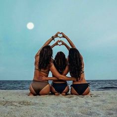 three women in bikinis sitting on the beach making a heart shape with their hands