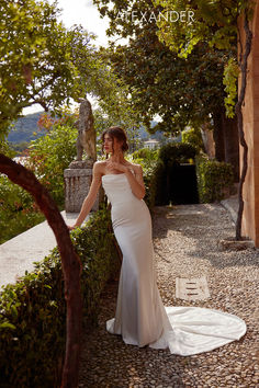 a woman in a white wedding dress standing on a stone path next to some trees