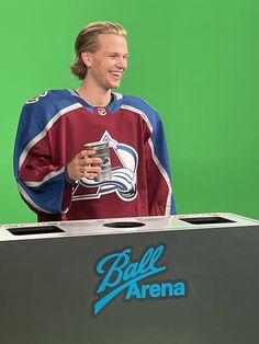 a man standing in front of a green screen holding a drink and wearing a hockey jersey