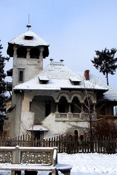 an old house with snow on the ground and a wooden bench in front of it