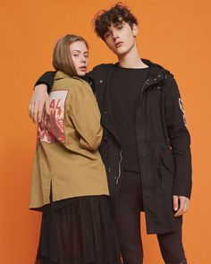 two young people standing next to each other in front of an orange wall, one wearing a black jacket