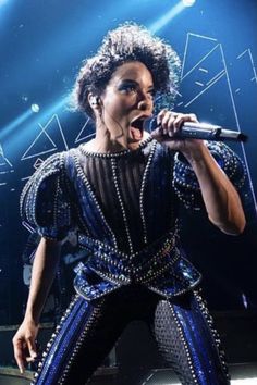 a woman in blue and black outfit holding a microphone on stage with lights behind her