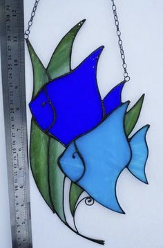 a blue glass fish hanging from a chain on a white wall next to a ruler