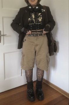 A mid-sized person/girl wearing a    black cropped graphic shirt, wide brown cargo shorts, black skull headphones, black cobweb tights, platform dr. Martens and multiple dark green and gray crystal rings and bracelets. 		The person is also wearing multiple silver necklaces. Masc Goth Outfits Plus Size, Fairycore Outfit Masc, Cottage Punk Aesthetic, Masc Goth Outfits Summer, Plus Size Alt Fashion Masc, Goblincore Masc Outfit