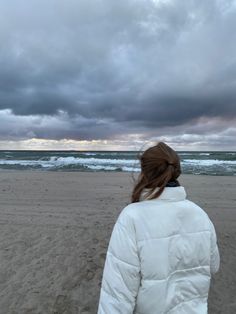 a woman standing on top of a sandy beach next to the ocean under a cloudy sky