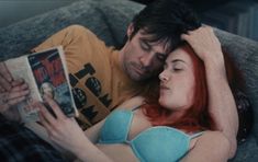 a man and woman laying on top of a couch reading a book while looking at each other