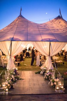 a wedding tent with white drapes and candles on the ground at night, filled with people