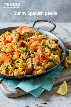 a skillet filled with rice and vegetables on top of a wooden table next to lemon wedges