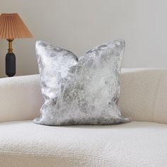 a silver pillow sitting on top of a white couch next to a light colored lamp