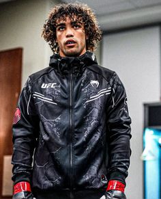 a man with curly hair wearing a black jacket and red gloves standing in front of a door